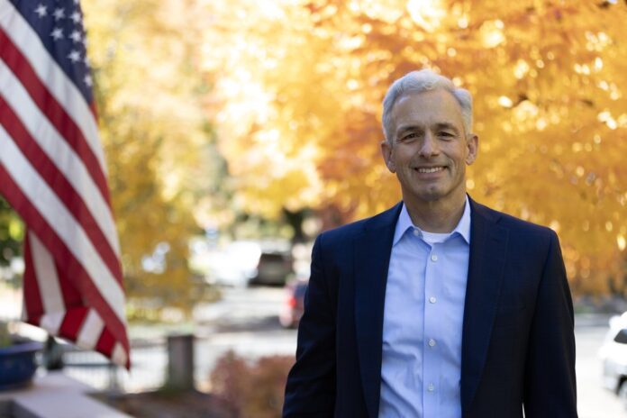 John Walsh, the victor in Denver’s District Attorney primary, stands outside in a blue suit jacket with an American flag and trees with fall leaves behind him.