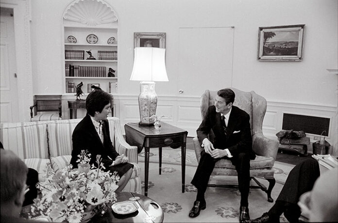 Anne Gorsuch, in formal clothing, sits on a couch in the Oval Office, next to former President Ronald Reagan, who sits in a chair.