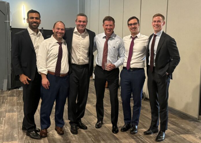 Giovanni Sanchez, Nosson Knobloch, Glen Summers, John Hughes, Taylor Kelson and Luke Beasley smile for a photo in various states of formal wear in an unadorned meeting room.