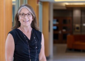 Lori Cowan smiles at the camera in a black dress, with a couch and office space out of focus behind her.