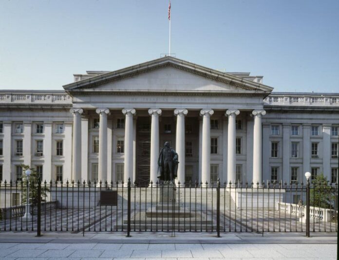 A view of the front of the U.S. Department of Treasury Building in Washington D.C. A short fence sits in the foreground in front of a statute. The building is white, with columns and a classical architectural style.