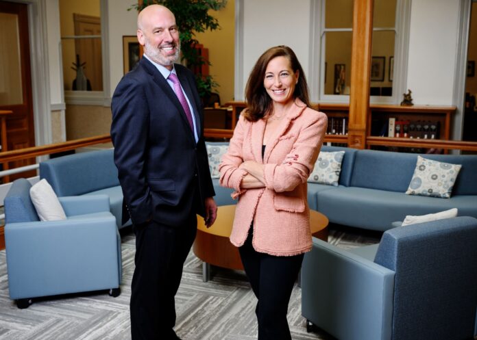 Cameron Garrison and Jennifer Johnson stand next to each other in the welcome area of an office. Garrison wears a navy suit and Johnson wears a tan jacket. In the background are couches and short bookshelves.