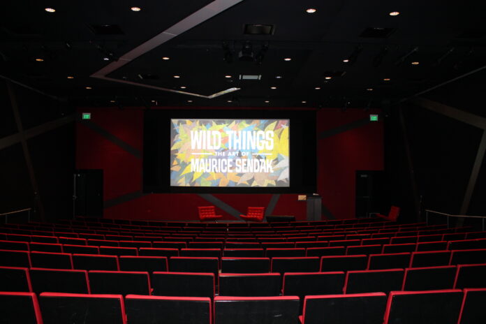 An empty auditorium in the basement of the Denver Art Museum sits ready for an art law CLE titled Wild Things: the Art of Maurice Sendak. A colorful cover of a slide deck is pulled up on a big screen with a podium to the right.