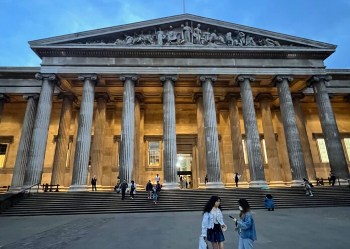 The front entrance to the British Museum, with a colonnade backlit by lights and a stone sculpture at the top of the colonnade.